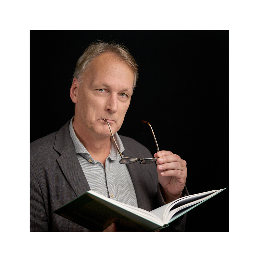 Fotografi av Fredrik Nilsson som håller upp en bok med ena handen och håller i sina glasögon med den andra.