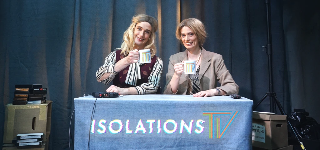 Föreställningsbild. De två programvärdarna i Isolations-TV sitter bakom ett bord med lyfta kaffekoppar och ler mot kameran.